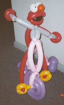 Elmo balloon character riding a purple and yellow tricycle